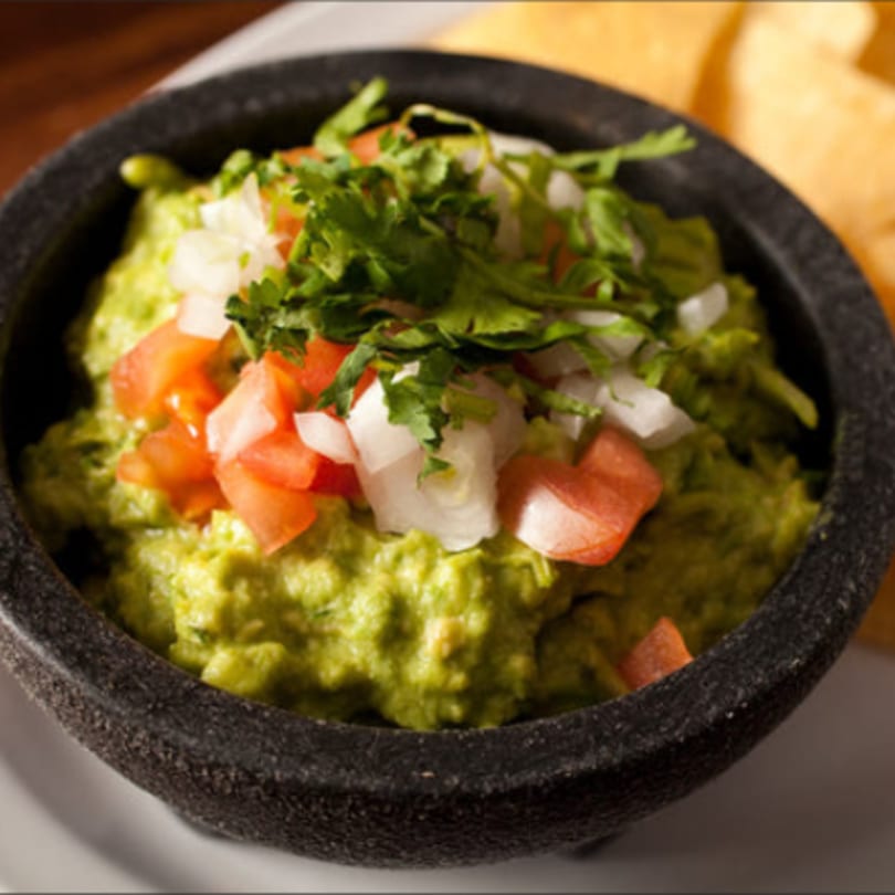 Guacamole in a dark stone bowl, topped with diced tomatoes, onions, and cilantro, with tortilla chips in the background.