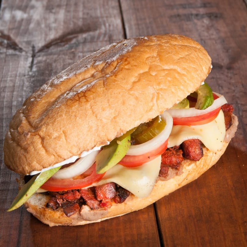 A large, crusty sandwich filled with meat, cheese, avocado, tomato, onion, and jalapeños on a wooden surface.